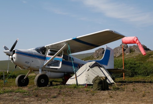 A small airplane parked next to a tent in a grassy area, with rocky hills in the background and a windsock nearby.