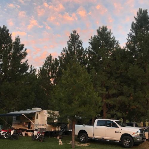 A campsite surrounded by tall trees, with a camper and a pickup truck under a colorful sunset sky.