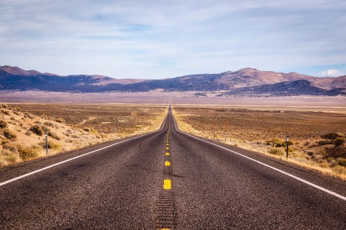 A long, straight road stretches into the distance, flanked by arid land and mountains under a clear sky.