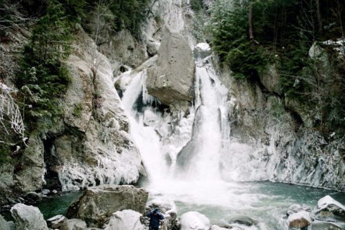 A frozen waterfall cascades into a rocky pool, surrounded by snow-covered trees and rugged cliffs.