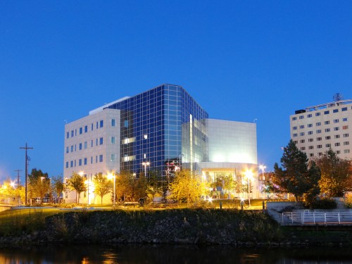 Modern building with glass facade, illuminated at dusk, alongside a river with trees and streetlights.