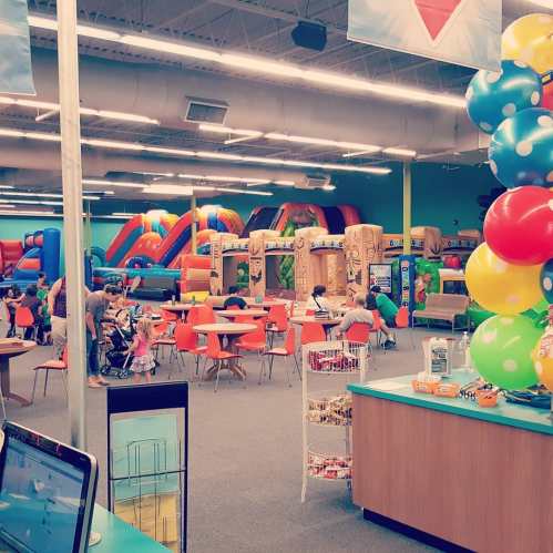 A colorful indoor play area with inflatable structures, tables, and children playing, decorated with balloons.