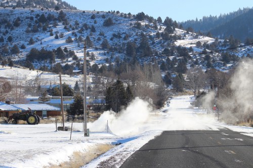 A snowy road with steam rising, surrounded by hills and trees, and a tractor in the background.