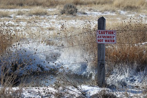 A sign warns of extremely hot water near a frosty landscape with dry grass and a small stream.