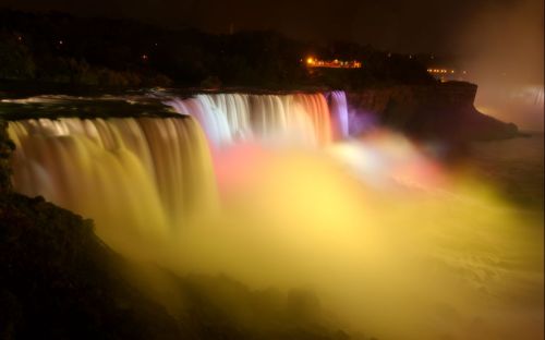 A vibrant waterfall illuminated by colorful lights at night, surrounded by mist and a dark sky.