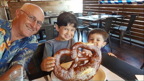 A smiling man and two boys hold a large pretzel at a restaurant, with wooden decor in the background.