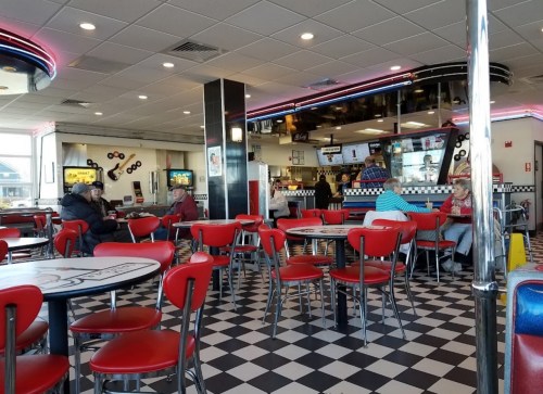 A retro diner interior with checkered floors, red chairs, and customers seated at tables, enjoying their meals.