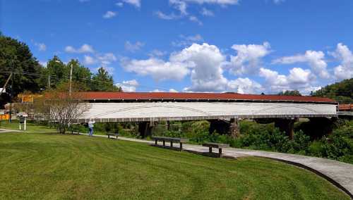 A covered bridge spans a river, surrounded by green grass and trees under a blue sky with fluffy clouds.