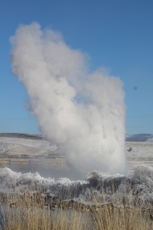 A plume of steam rises against a clear blue sky, surrounded by frosty grass and distant hills.