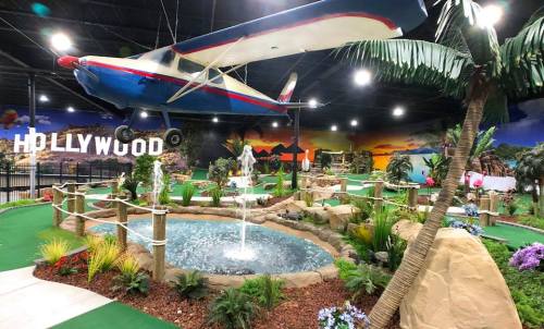 A vibrant indoor mini-golf course featuring a suspended airplane, palm trees, a pond, and scenic Hollywood-themed decor.