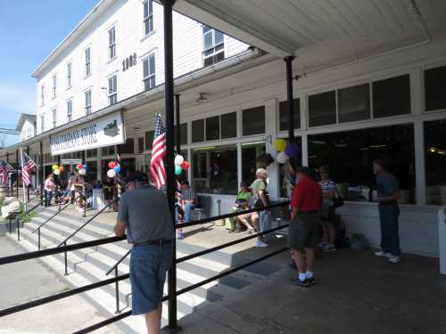 A crowd gathers outside a store with balloons and American flags, enjoying a festive atmosphere on a sunny day.