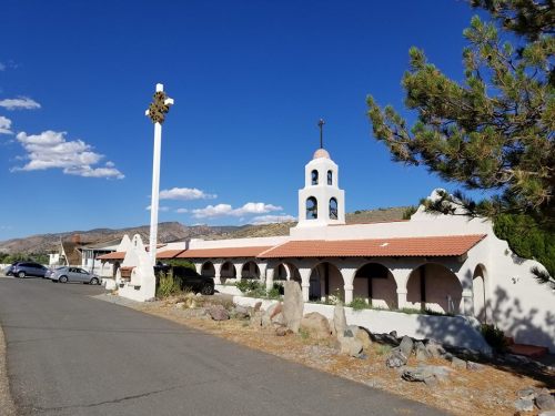 A white building with a bell tower and arched windows, set against a blue sky with scattered clouds and mountains in the background.