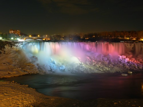 A nighttime view of Niagara Falls illuminated in colorful lights, with icy surroundings and a calm river below.