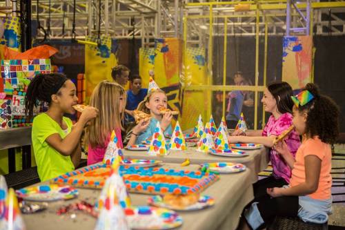A group of children at a birthday party, wearing party hats and enjoying pizza and colorful cake in a fun indoor setting.