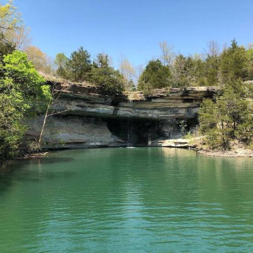 A serene natural landscape featuring a clear blue-green lake, lush greenery, and a rocky cliff with a waterfall.