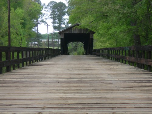 A wooden covered bridge stretches over a path surrounded by lush green trees.