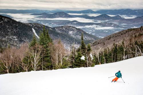 A skier descends a snowy slope, surrounded by mountains and a misty valley below.