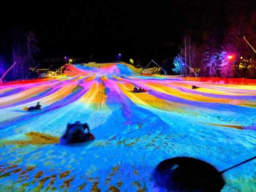 Colorful lights illuminate a snowy hill as people sled down the slopes at night.