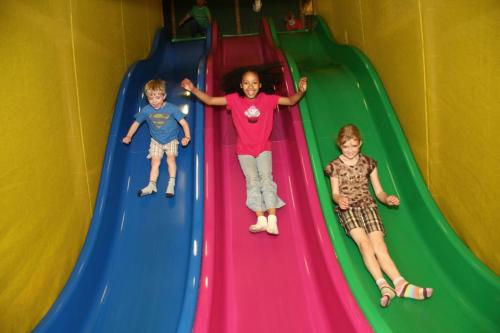 Three children slide down colorful slides in a play area, smiling and enjoying their time together.