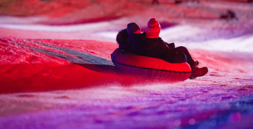 Two people sit on a snow tube, sliding down a brightly lit snowy slope at night.