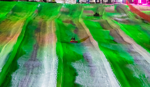 Two people sledding down a brightly lit, colorful slope at night, surrounded by vibrant green and purple lights.