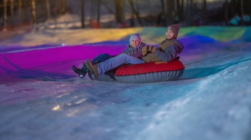 A man and a child sit on a red snow tube, sliding down a snowy hill illuminated by colorful lights at night.