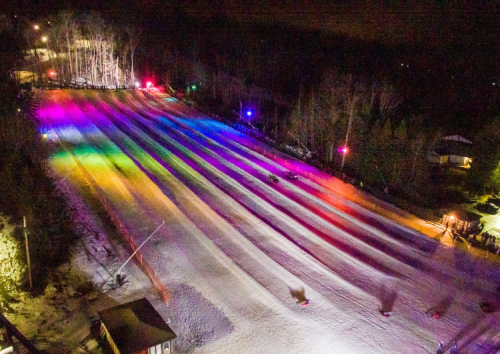 A snowy hill at night, illuminated by colorful lights, with people sledding down the slopes.