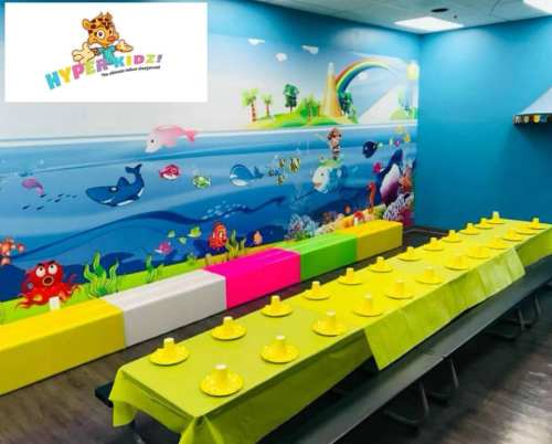 A colorful party room with a mural of underwater scenes, featuring a long table set with yellow cups and bright seating.