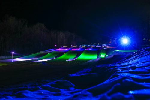 A snowy slope illuminated by colorful lights at night, with gentle hills and a bright spotlight in the background.