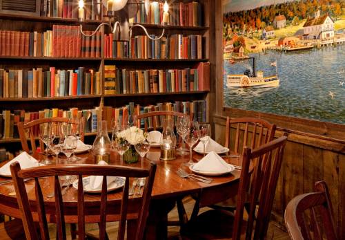 A cozy dining area with a wooden table set for dinner, surrounded by bookshelves and a scenic mural of a waterfront.
