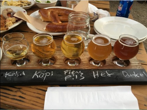 A wooden flight board with five different beer samples, alongside plates of food and a drink in the background.