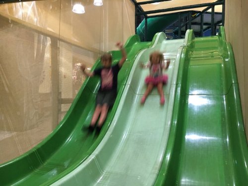 Two children sliding down green slides in an indoor play area, with excitement on their faces.