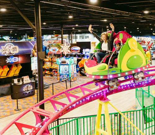 A colorful indoor roller coaster with two excited riders, surrounded by arcade games and bright lights.