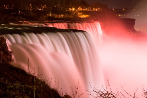 A nighttime view of a waterfall illuminated in soft pink light, with mist rising and city lights in the background.