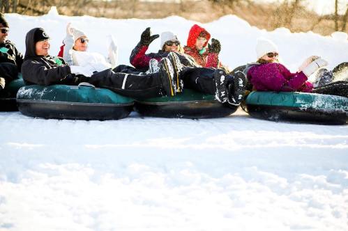 A group of people enjoying snow tubing, laughing and waving while sitting on inflatable tubes in a snowy landscape.