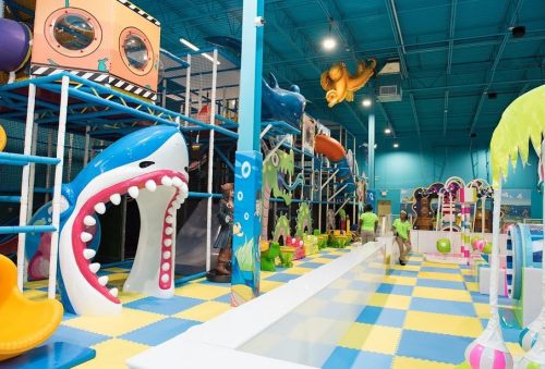 Colorful indoor play area with slides, climbing structures, and a large shark feature, filled with children and playful decor.