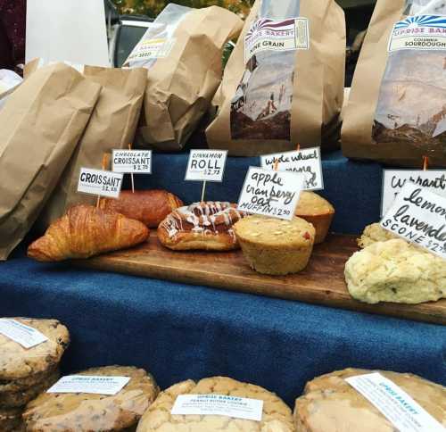 A display of baked goods including croissants, rolls, and muffins, with labeled paper bags in the background.