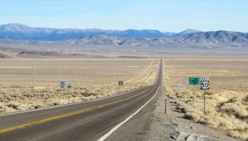 A long, straight road stretches through a vast, arid landscape with mountains in the distance under a clear blue sky.