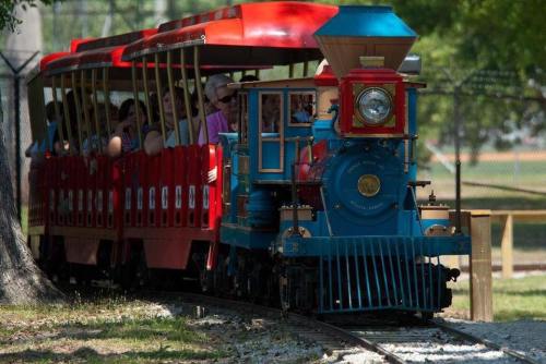 A colorful miniature train with passengers rides along a track in a park, surrounded by trees.