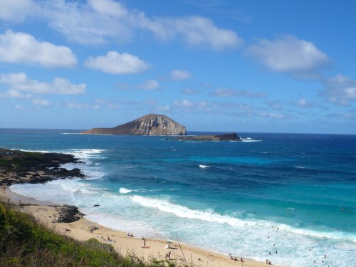 A scenic view of a beach with waves, a rocky coastline, and a distant island under a blue sky with fluffy clouds.