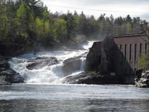 A waterfall cascades over rocks near a dam, surrounded by trees and a cloudy sky.