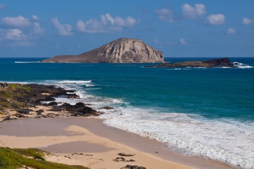 A scenic beach with golden sand, rocky shoreline, and a distant island under a blue sky with fluffy clouds.