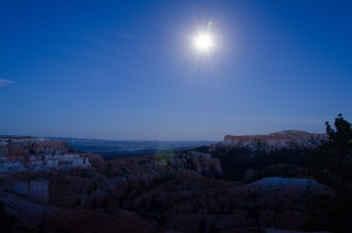 A serene night scene of Bryce Canyon under a bright full moon, illuminating the rocky landscape and distant hills.