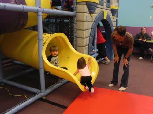 Two children play on a yellow slide in a colorful indoor playground, while an adult watches and encourages them.