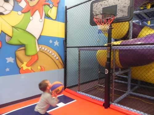 A child shoots a basketball at a hoop in a colorful indoor play area with a slide in the background.