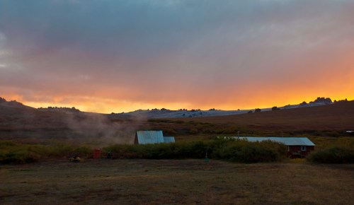 A serene landscape at sunset, featuring a red barn, rolling hills, and a colorful sky with clouds.