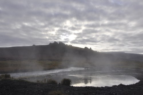 A misty landscape with a steaming body of water, surrounded by hills under a cloudy sky.
