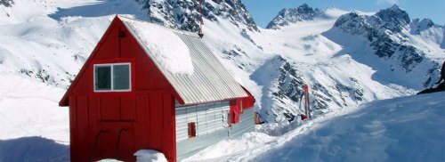 A red cabin surrounded by snow-covered mountains under a clear blue sky.