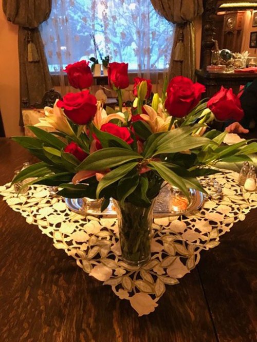 A vase of red roses and lilies on a lace tablecloth, with a cozy indoor setting and snowy window in the background.