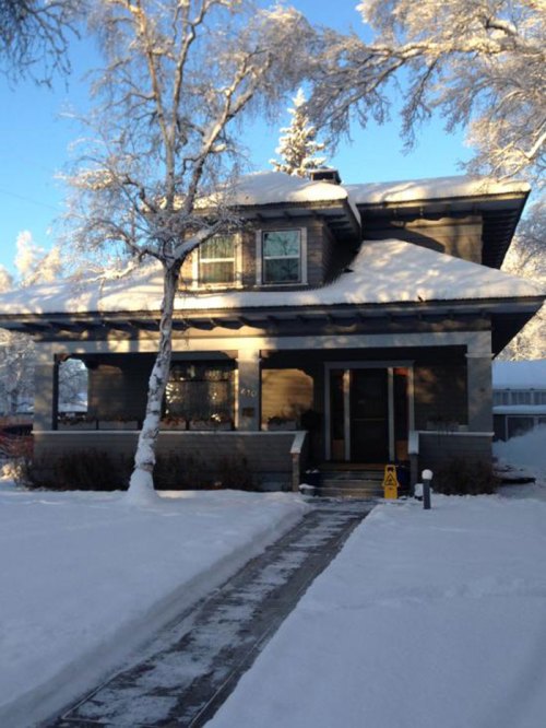 A snow-covered house with a dark exterior, surrounded by trees and a clear blue sky. Snow blankets the ground.